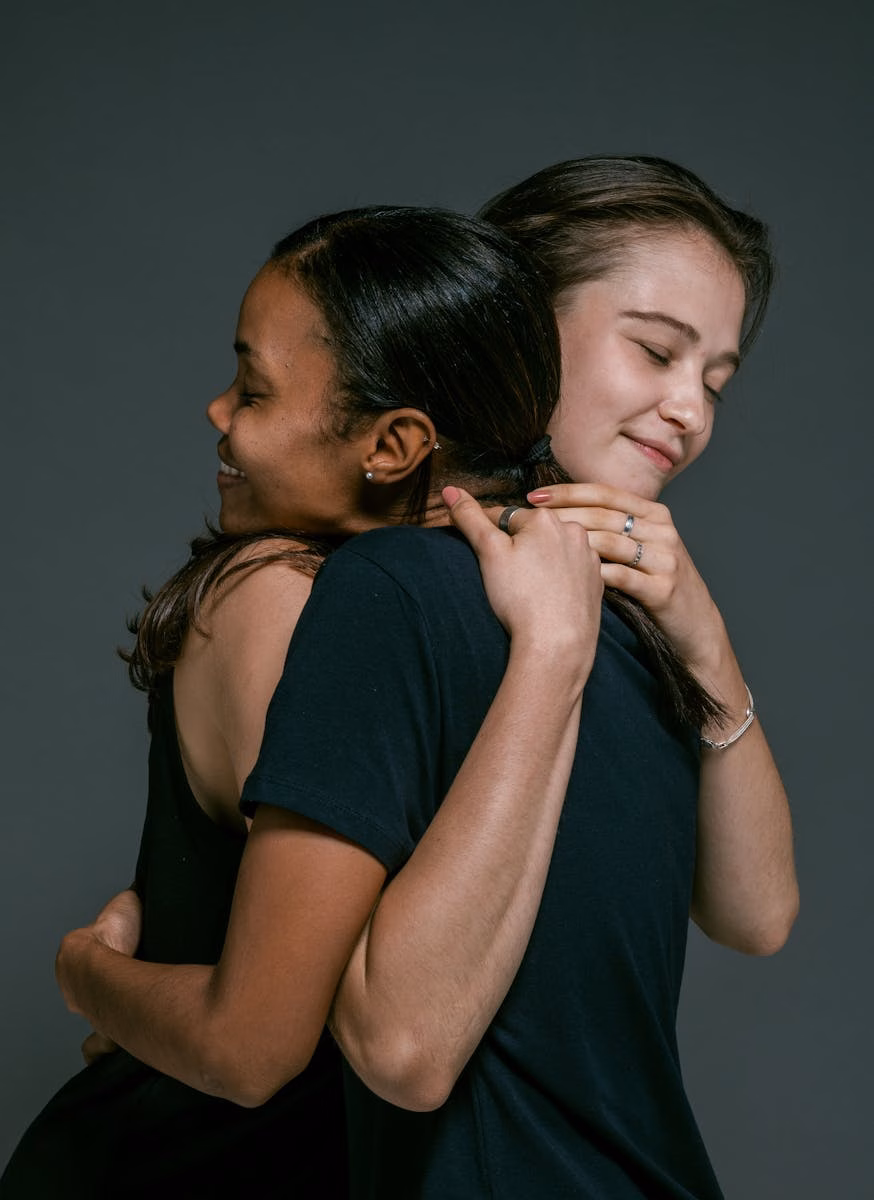 Two women of different ethnicities share a warm hug, symbolizing love and friendship against a gray backdrop.